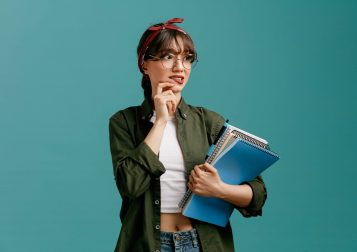 anxious young student girl wearing bandana glasses holding large note pads with pen looking at side touching chin biting lip isolated on blue background